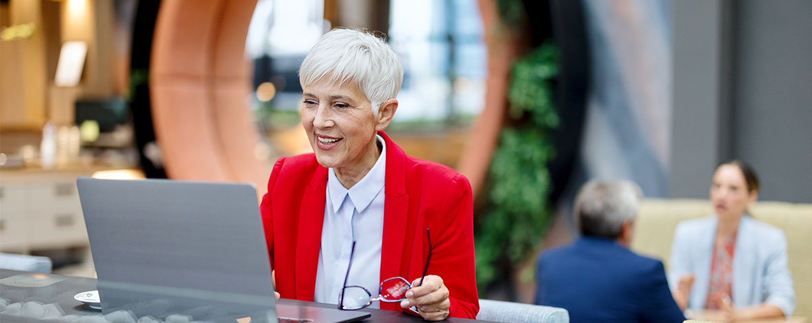 Une dame âgée en costume rouge qui consulte son ordinateur dans un open space